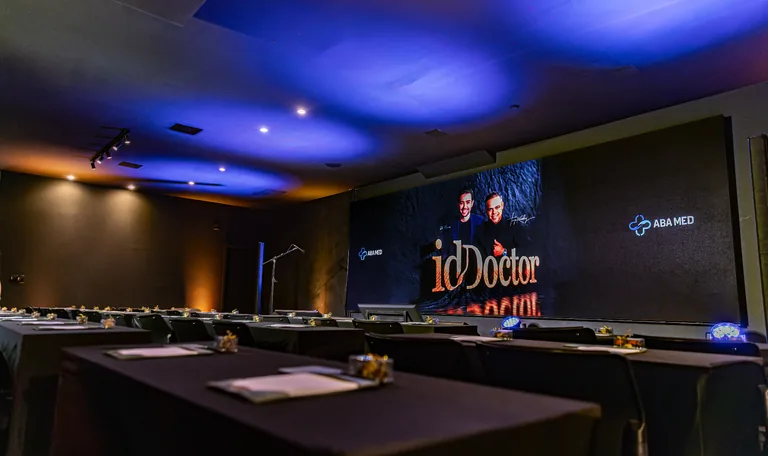 Auditório da Casa do Relacional pronto para evento, com painel LED idDoctor e ABA MED ao fundo, iluminação cênica âmbar e mesas em formato escolar