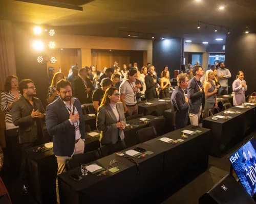 Plateia de médicos e dentistas em pé com a mão no peito durante momento solene do evento idDoctor